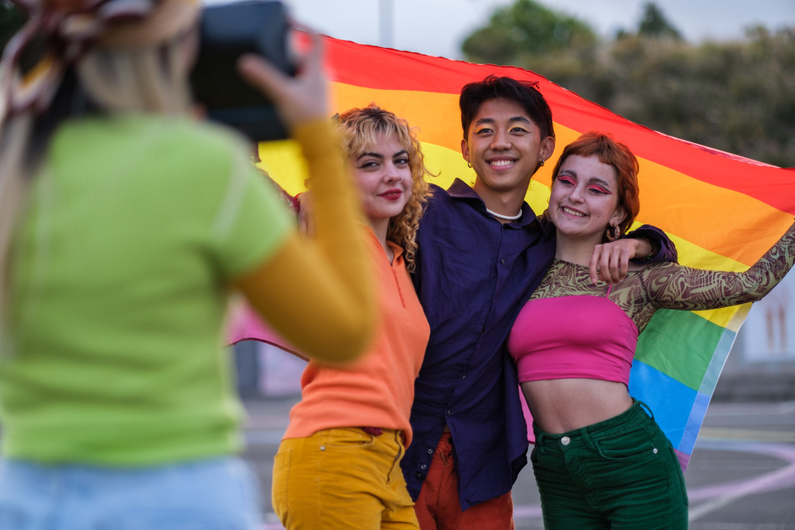 Three friends pose happily under a rainbow Pride flag while someone takes their picture outdoors.