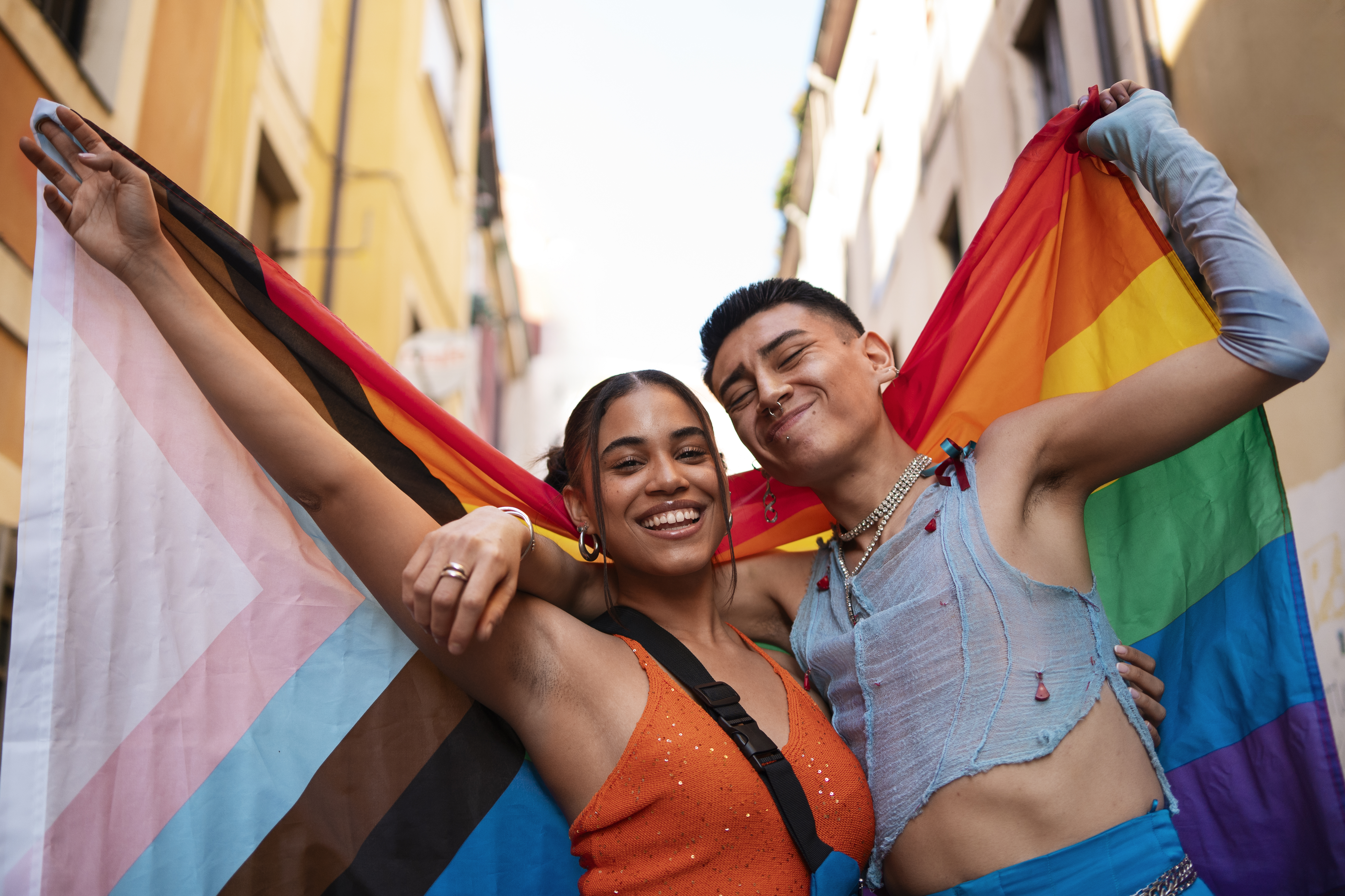 Two friends celebrate Pride outdoors, smiling and holding a Progress Pride flag together.