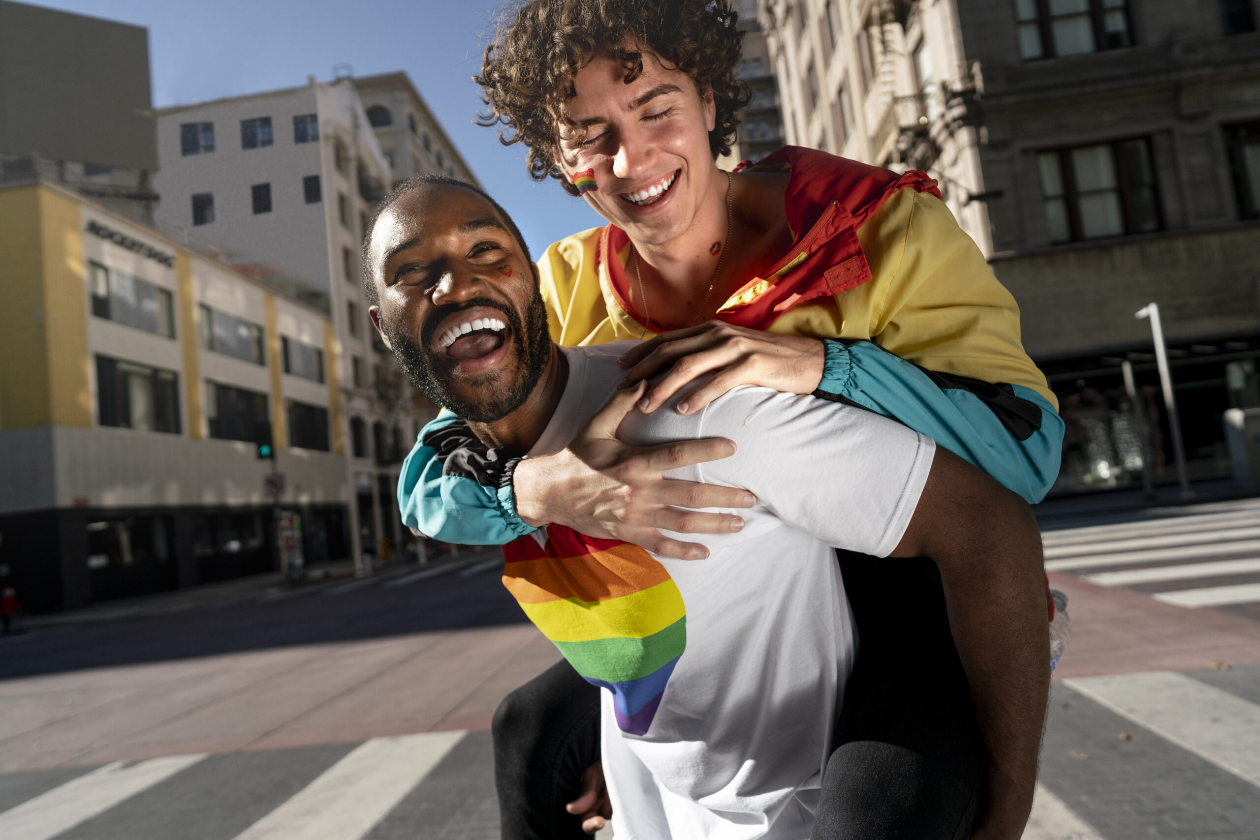 Two joyful men celebrate Pride outdoors, one giving the other a piggyback ride and both laughing.